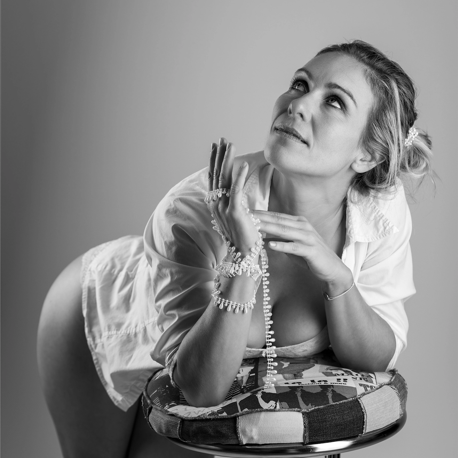 Schwarz-Weiß-Portraitfotografie im Studio. Model lehnt auf Barhocker und blickt nach oben. Um ihre rechte Hand hat sie eine Perlenkette.